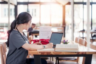Female Asian-American nursing student wearing gray scrubs sits in a cafe doing her coursework on her laptop surrounded by books and notes.