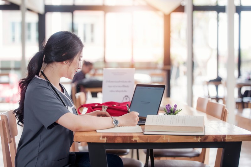 Female Asian-American nursing student wearing gray scrubs sits in a cafe doing her coursework on her laptop surrounded by books and notes.