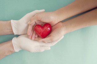 A pair of latex gloved hands holding bare hands that hold a red heart