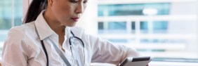 Asian-American female healthcare provider wearing a stethoscope and sitting in the window of a hospital reviewing her patients' charts on a tablet.