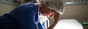 Stressed female nurse in PPE leans on bed and rubs forehead
