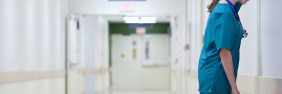 Stressed CRNA in blue scrubs holding a pink folder and leaning head against wall of white hospital hallway.