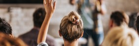 Looking at the back of a young blonde woman raising her hand in the middle of a crowded seminar.