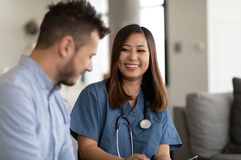 A young doctor of Asian descent and a young Caucasian patient are indoors at the man's home. The doctor is talking to the man while holding a tablet computer.