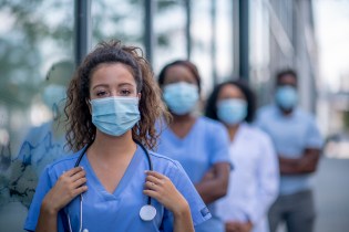 Masked female nurse practitioner standing outside facing camera with row of NPs standing behind her.