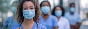 Masked female nurse practitioner standing outside facing camera with row of NPs standing behind her.
