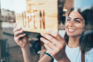 Young white woman with dark hair putting up a We're Open sign in her shop's front door