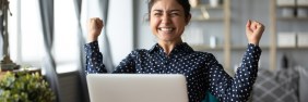 Young woman of color raising her arms in excitement while sitting in front of laptop.