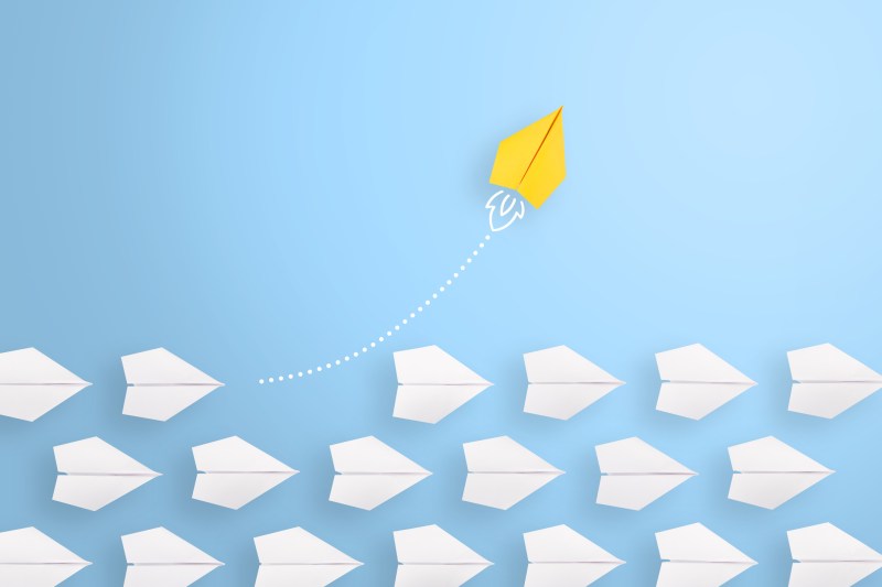 One yellow paper airplane breaking away from the pack of white paper airplanes flying against a baby blue background.