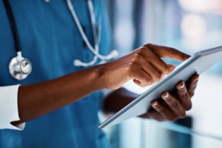 Lower half of African-American man wearing blue scrubs and a stethoscope scrolling through a document on a tablet.