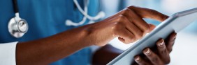 Lower half of African-American man wearing blue scrubs and a stethoscope scrolling through a document on a tablet.