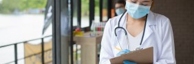 Young Asian-American female nurse wearing mask, white lab coat, and stethoscope taking notes on a clipboard in the middle of a hallway.