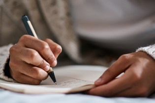 Woman in a beige sweater writing a draft of her cover letter in her notebook.