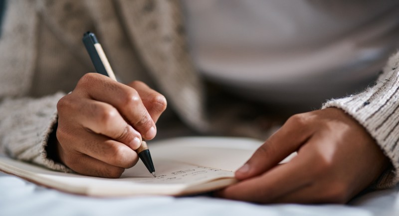 Woman in a beige sweater writing a draft of her cover letter in her notebook.