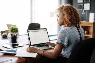 Young African-American woman sitting at a desk writing in a notebook while doing research on her laptop.