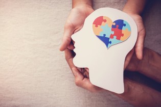 Two pairs of hands holding a white cardstock cutout of a human head with a colorful heart-shaped jigsaw puzzle in the brain.