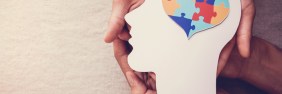 Two pairs of hands holding a white cardstock cutout of a human head with a colorful heart-shaped jigsaw puzzle in the brain.