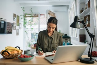 Young woman sitting at desk writing in notebook with laptop open in front of her.