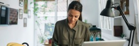 Young woman sitting at desk writing in notebook with laptop open in front of her.