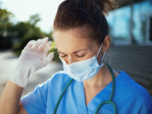 female nurse in mask holding her hand to her head looking stressed | rn licensing board complaint