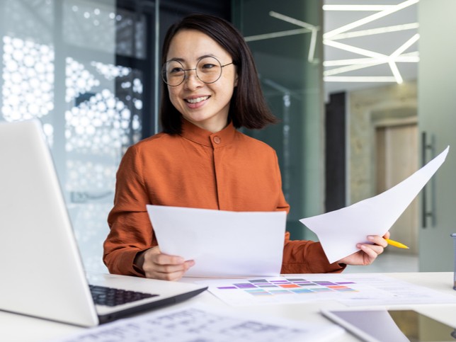 Woman in orange holding papers at her desk while smiling | Errors and Omissions Insurance Costs